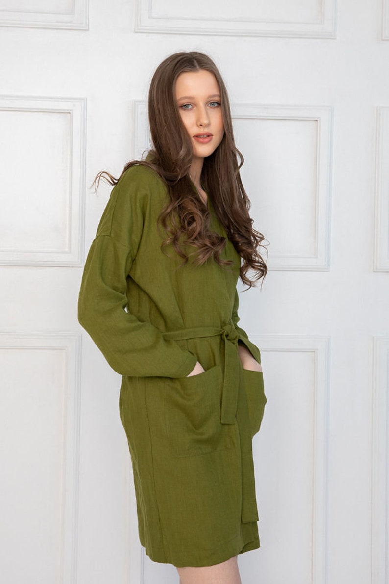 May include: A long-sleeved, olive green linen dress with a tie belt and two large pockets. The dress is made of a lightweight, breathable fabric and has a relaxed fit.