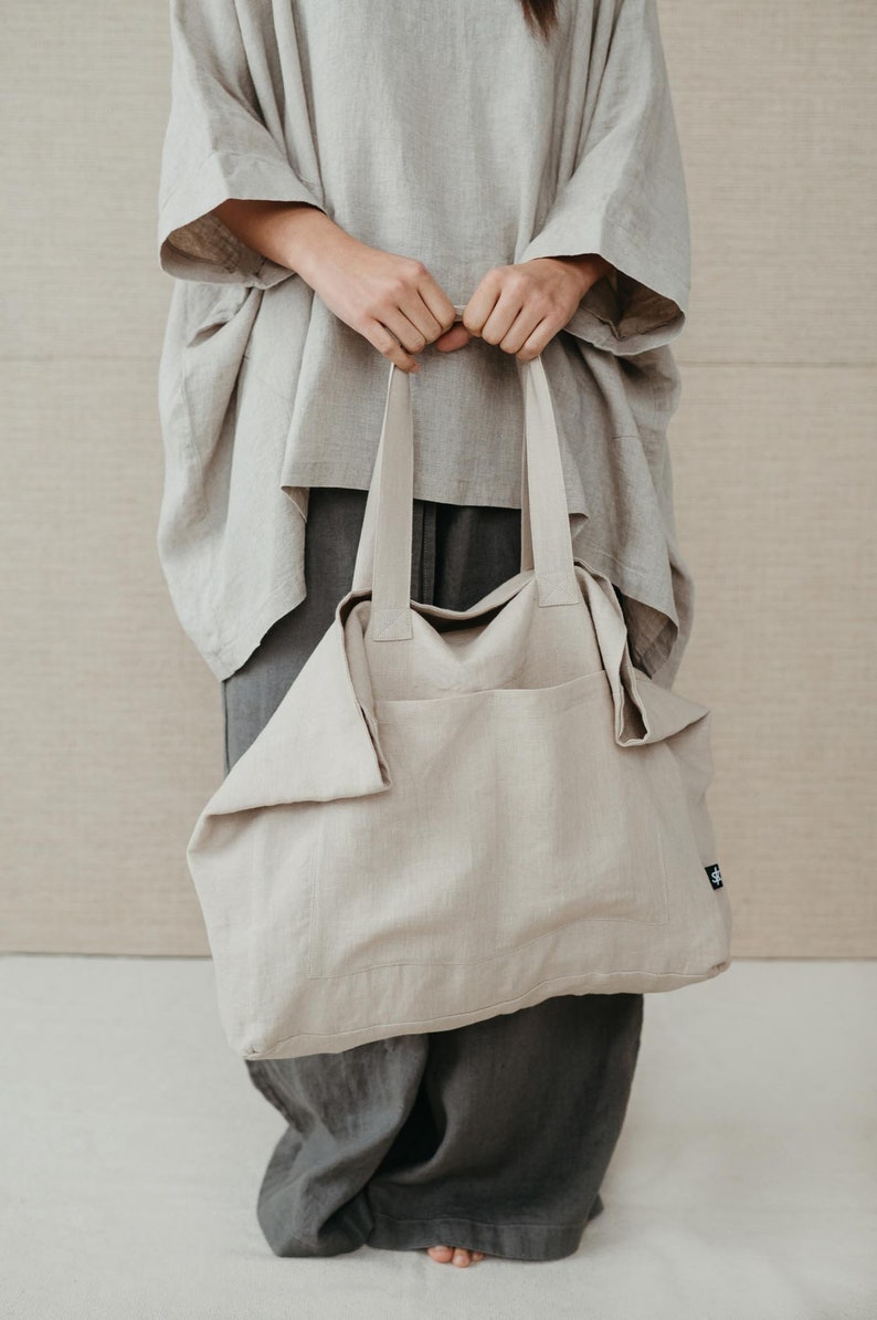 May include: A large, light beige linen tote bag with thick straps. The bag is folded over at the top and has a small black label on the side.