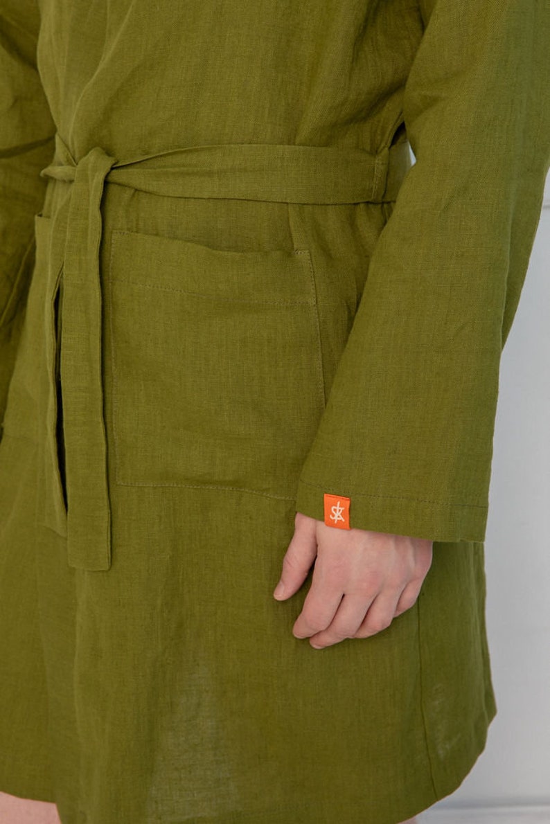 May include: A green linen robe with a belt and a pocket. The robe has a small orange label with the letters "SK" on the sleeve.