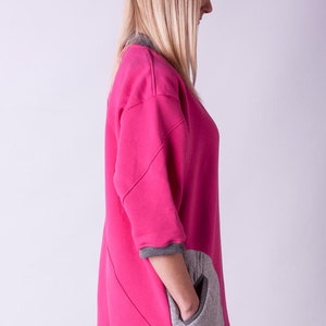 May include: A pink and gray sweatshirt with a large pocket on the side. The sweatshirt has a relaxed fit and a long, flowing design.