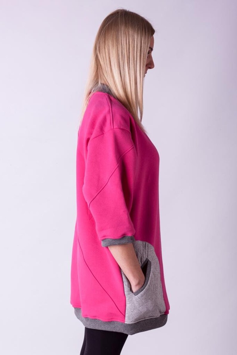 May include: A pink and gray sweatshirt with a large pocket on the side. The sweatshirt has a relaxed fit and a long, flowing design.