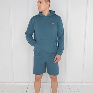 Puede incluir: Un conjunto de sudadera con capucha y pantalones cortos a juego de color azul turquesa. La sudadera tiene un pequeño logotipo bordado en el pecho izquierdo.