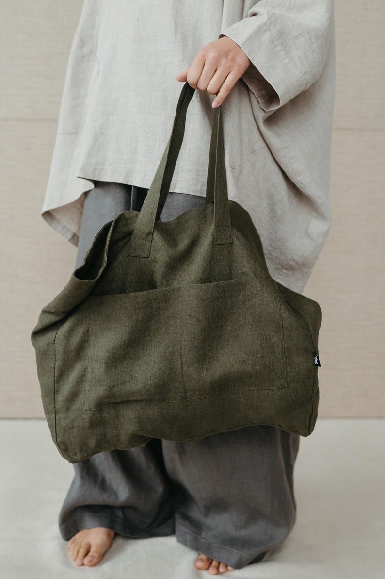 May include: A large, olive green linen tote bag with a single pocket on the front. The bag has thick, sturdy straps.