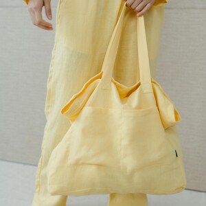 May include: A person wearing a yellow linen jumpsuit holds a large yellow linen tote bag with a single pocket.