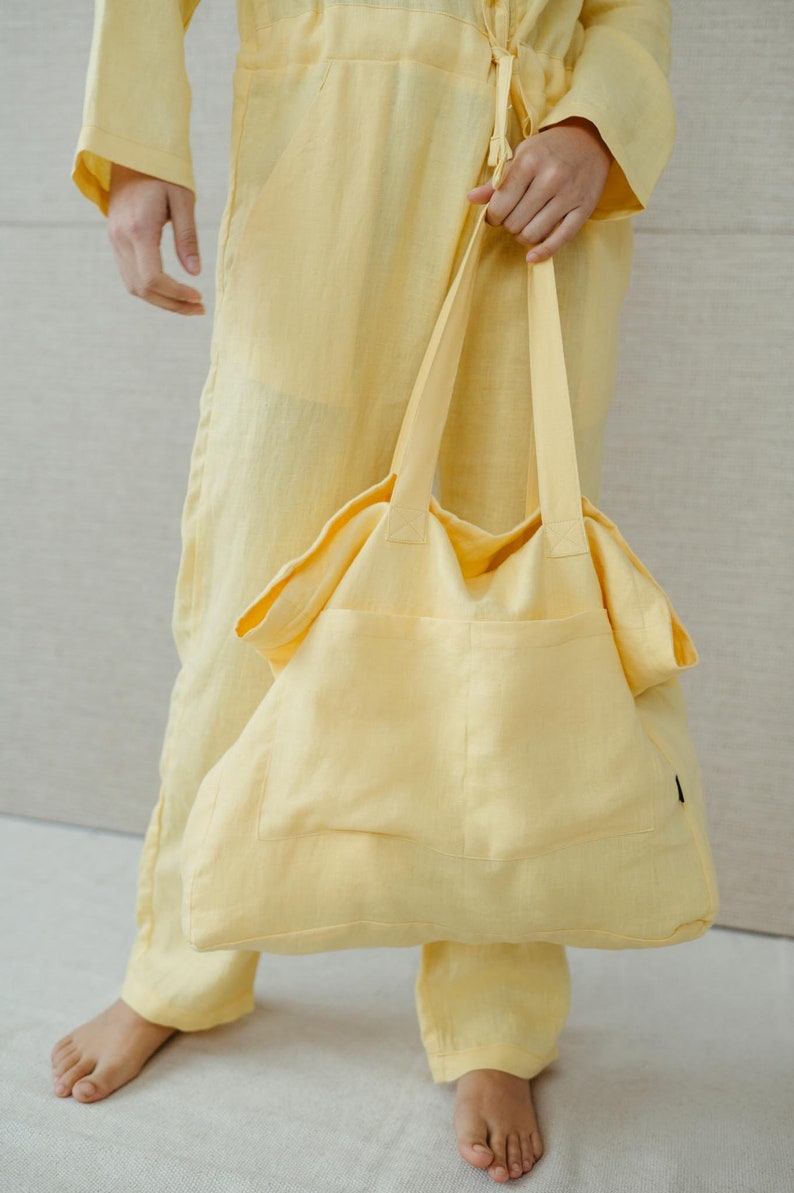 May include: A person wearing a yellow linen jumpsuit holds a large yellow linen tote bag with a single pocket.