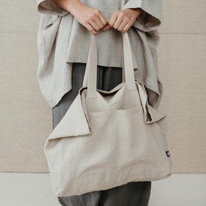 May include: A large, light beige linen tote bag with thick straps. The bag is folded over at the top and has a small black label on the side.