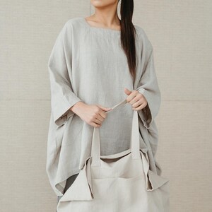 May include: A woman wearing a light gray linen top and gray pants is holding a large, off-white linen tote bag with a small black logo.