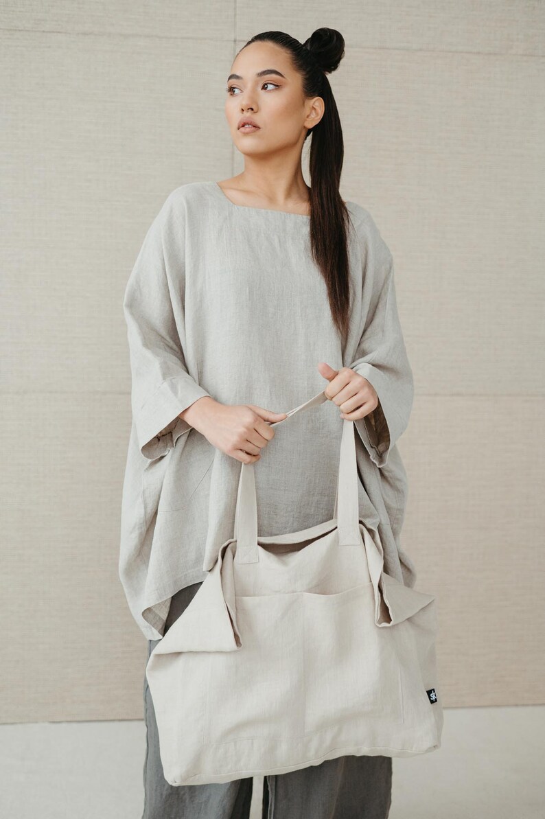 May include: A woman wearing a light gray linen top and gray pants is holding a large, off-white linen tote bag with a small black logo.
