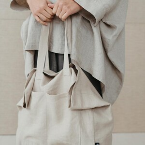 May include: A large, beige linen tote bag with a single pocket on the front. The bag has a small black label with a white logo on the bottom right corner.