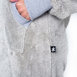 May include: A grey fleece jumpsuit with a pocket on the left side and a black label with the letters "SK" on the right side.