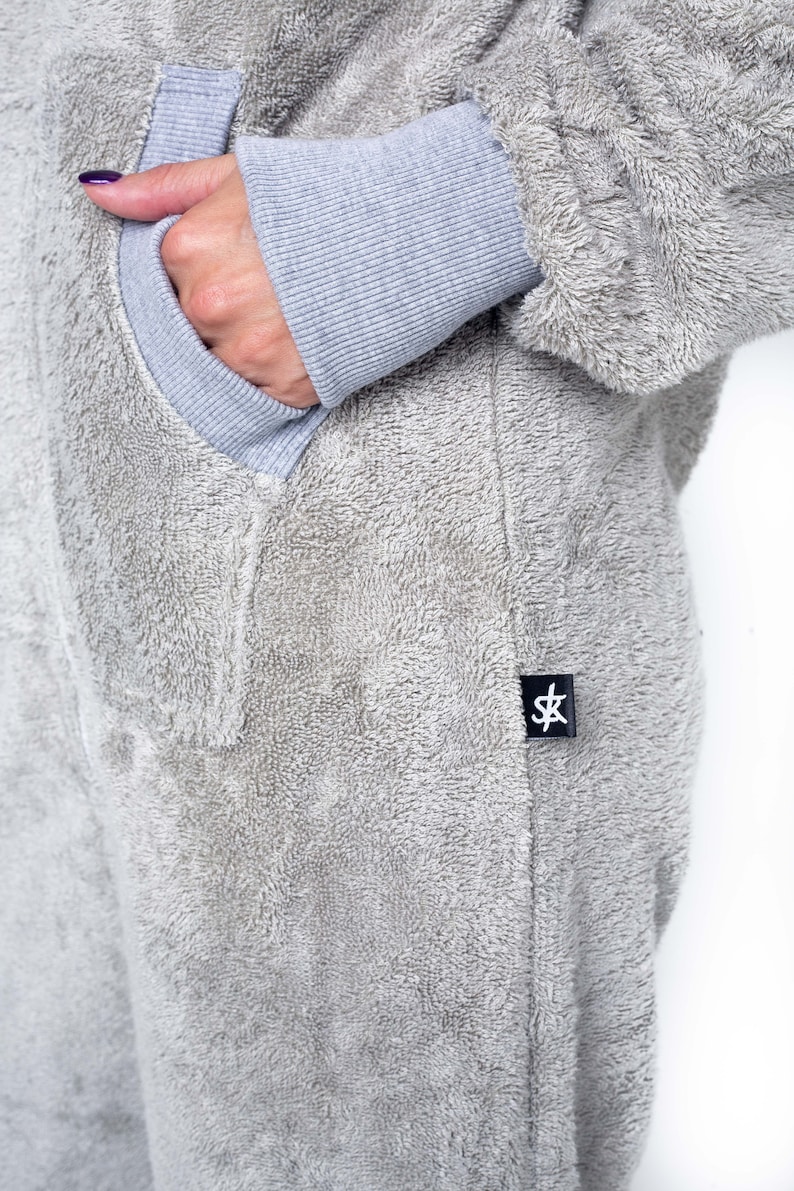 May include: A grey fleece jumpsuit with a pocket on the left side and a black label with the letters "SK" on the right side.