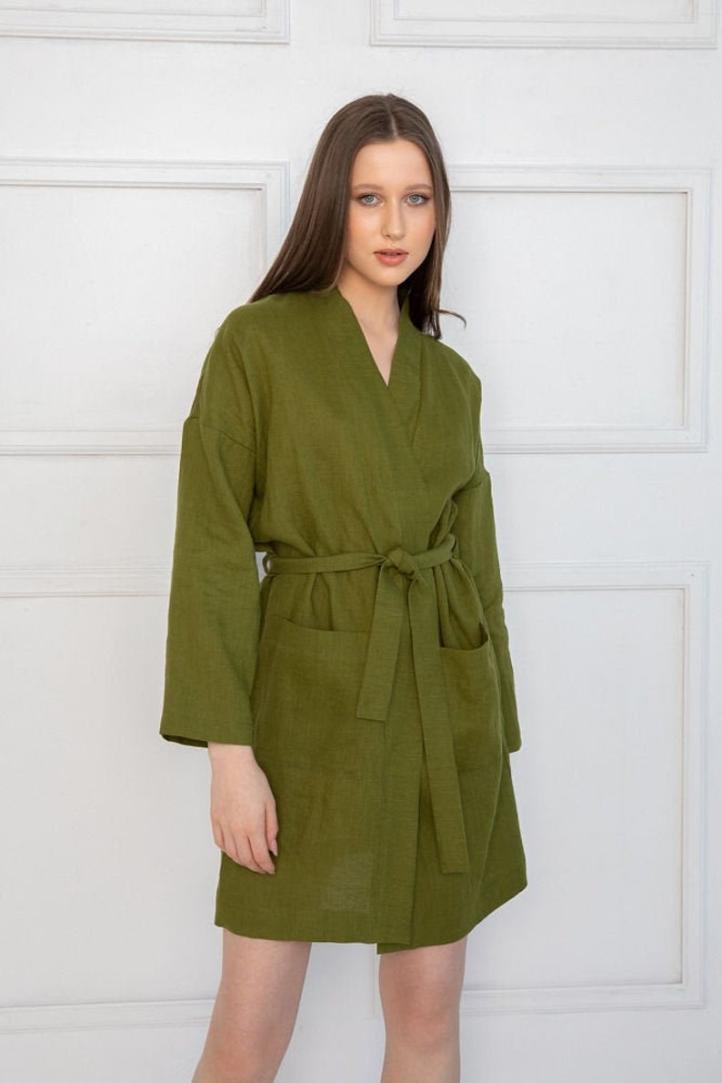 May include: A long-sleeved, olive green linen robe with a V-neck and a tie belt. The robe has two pockets and a relaxed fit.