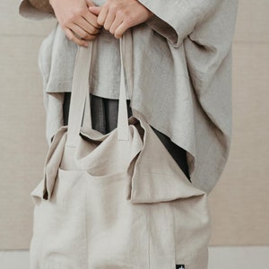 May include: A large, beige linen tote bag with a single pocket on the front. The bag has a black label with a white logo on the bottom right corner.
