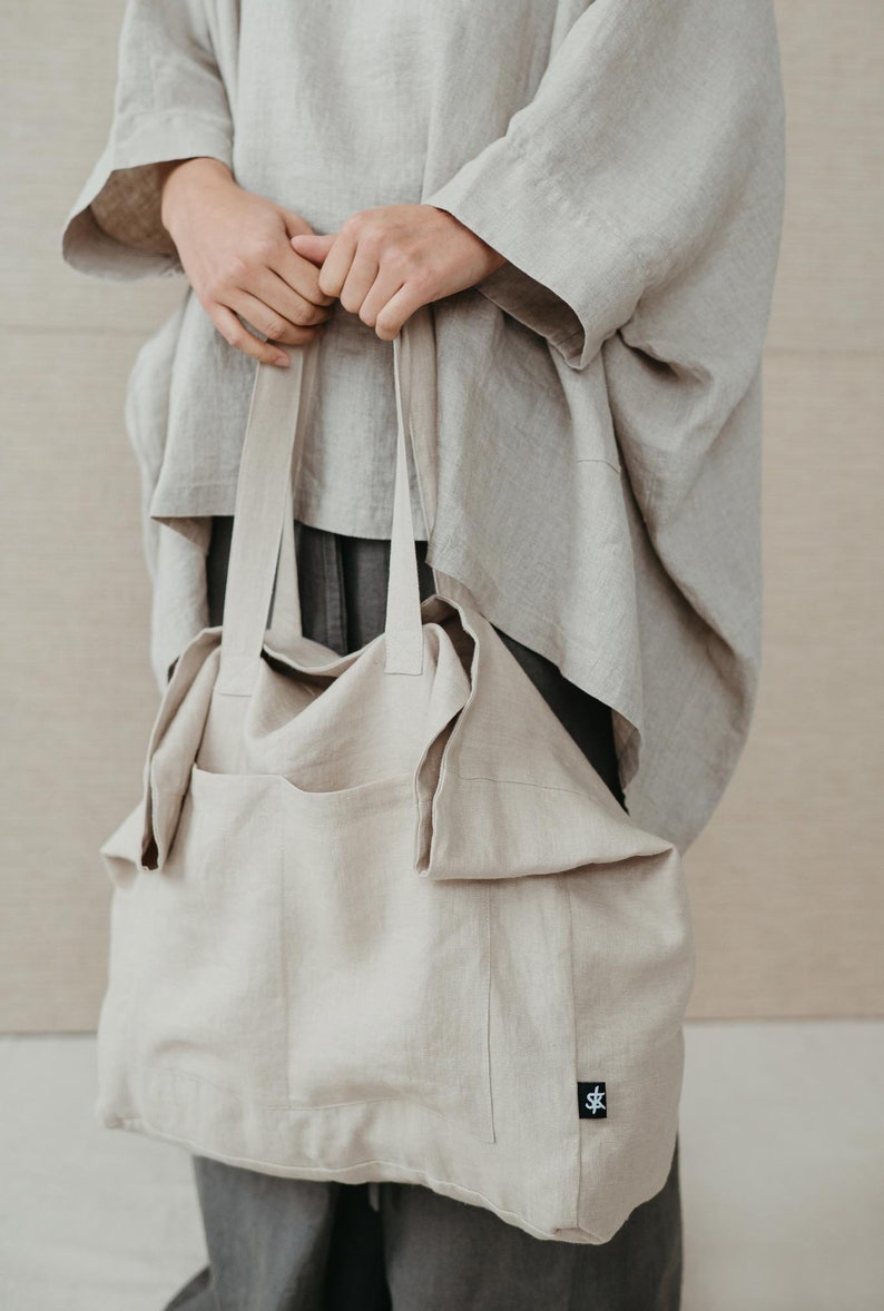 May include: A large, beige linen tote bag with a single pocket on the front. The bag has a black label with a white logo on the bottom right corner.