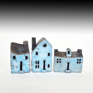 Op de afbeelding: Drie keramische miniatuurhuizen met een lichtblauwe glazuur en donkergrijze accenten. Elk huis heeft een uniek ontwerp, inclusief schoorstenen en ramen. De huizen zijn in een rij gerangschikt en tonen hun handgemaakte details.