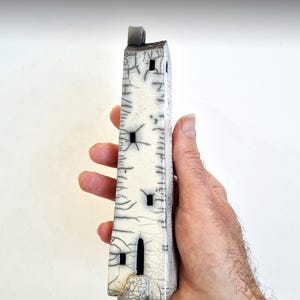May include: A small white ceramic tower with black windows and a cracked glaze. The tower is held in a person's hand.