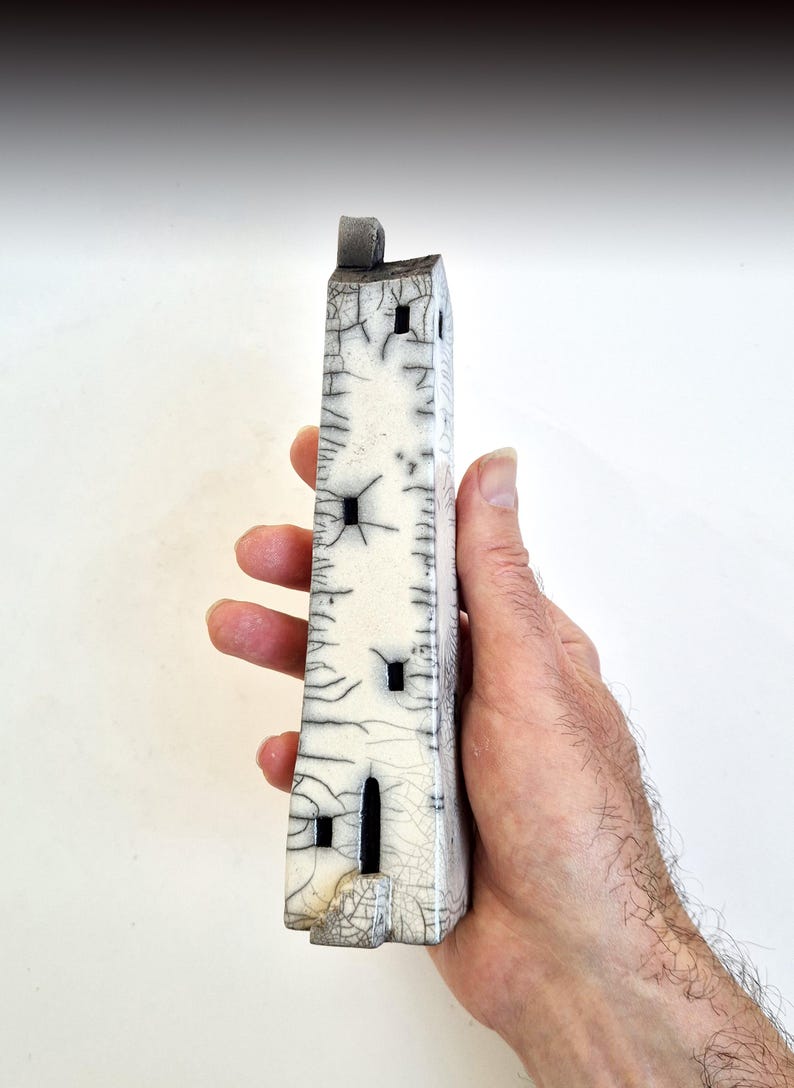 May include: A small white ceramic tower with black windows and a cracked glaze. The tower is held in a person's hand.
