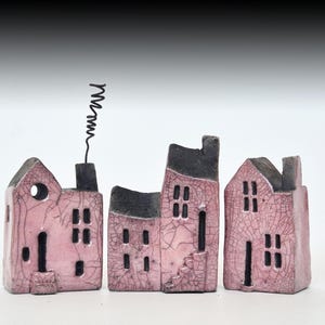 May include: Three miniature ceramic houses with a light pink crackle glaze and dark grey roofs. The structures feature rectangular windows, doorways, and a black wire chimney detail on the leftmost house. Each piece has a textured, weathered finish.