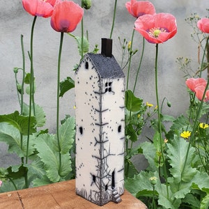 May include: A white ceramic house with black details and a small window, sitting on a wooden surface in front of a background of pink poppy flowers and green foliage.