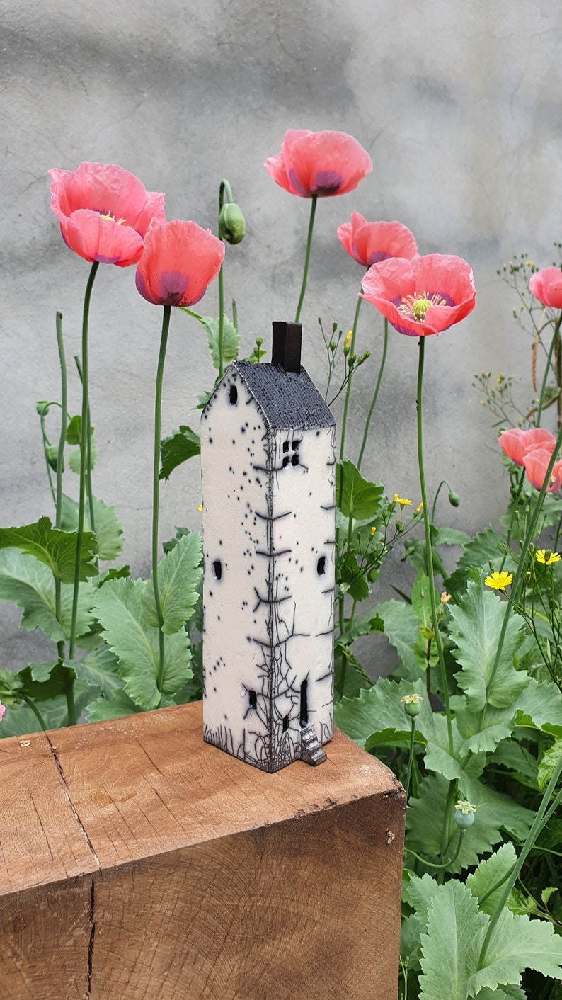 May include: A white ceramic house with black details and a small window, sitting on a wooden surface in front of a background of pink poppy flowers and green foliage.