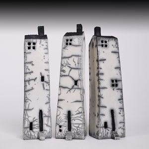 May include: Three white ceramic houses with black accents and crackled glaze. The houses are tall and narrow, with small windows and doors. They are arranged in a row on a white background.