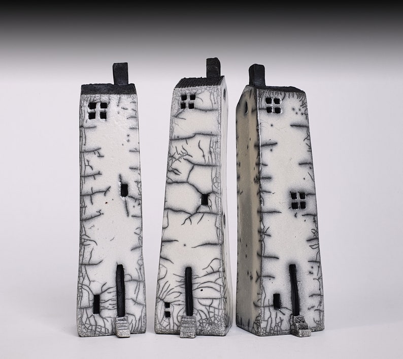 May include: Three white ceramic houses with black accents and crackled glaze. The houses are tall and narrow, with small windows and doors. They are arranged in a row on a white background.