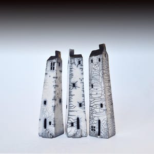 May include: Three white ceramic sculptures of houses with black details. The houses are tall and narrow, with a textured surface. They are arranged in a row, from left to right.