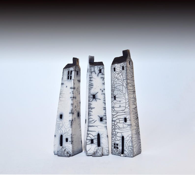 May include: Three white ceramic sculptures of houses with black details. The houses are tall and narrow, with a textured surface. They are arranged in a row, from left to right.