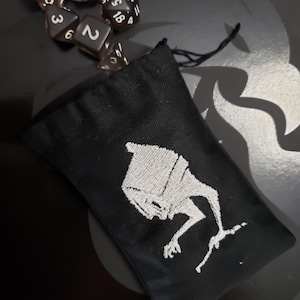 May include: Black drawstring bag with a white embroidered image of a creature with a large head and long arms. The creature is in a crouching position.