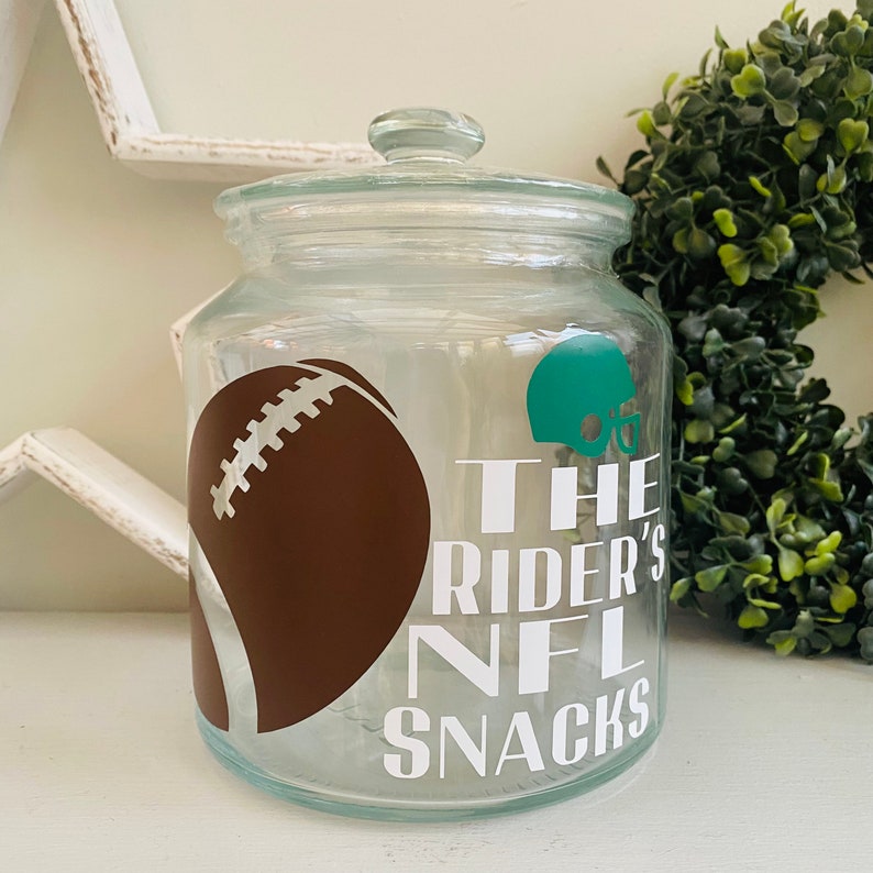 May include: A clear glass jar with a lid, decorated with a brown football, a green football helmet, and the text "The Rider's NFL Snacks".