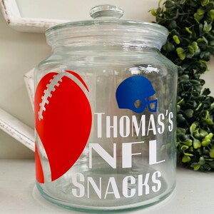 May include: Clear glass jar with a white lid. The jar has a red and white football and a blue football helmet with the text "Thomas's NFL Snacks" on the front.