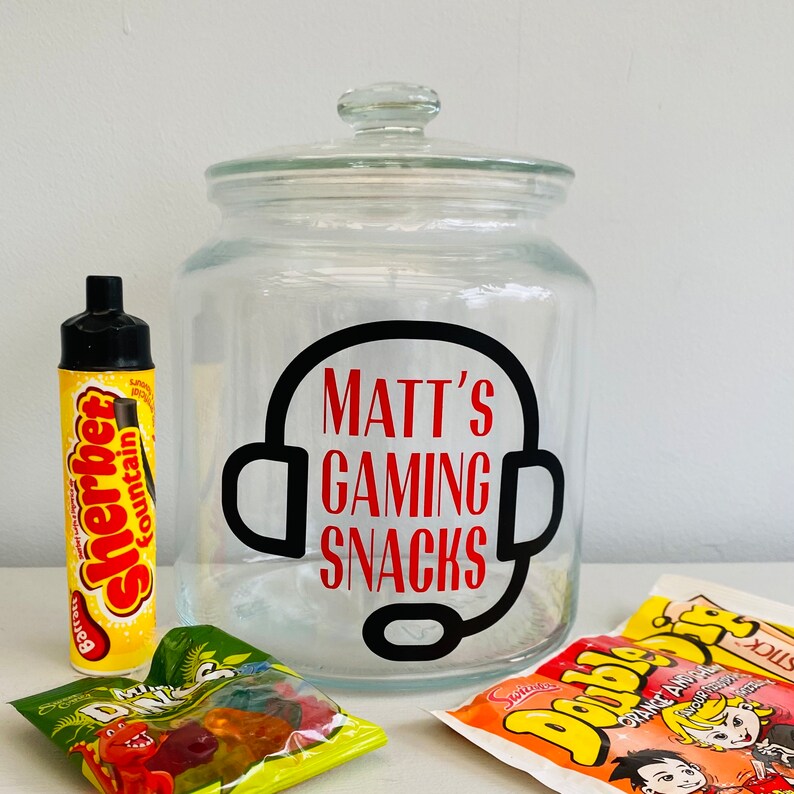 May include: A clear glass jar with a lid, labeled "Matt's Gaming Snacks" in red text, surrounded by a black headset graphic. The jar is surrounded by various snacks, including a yellow "Sherbet Fountain" tube and candy packets.