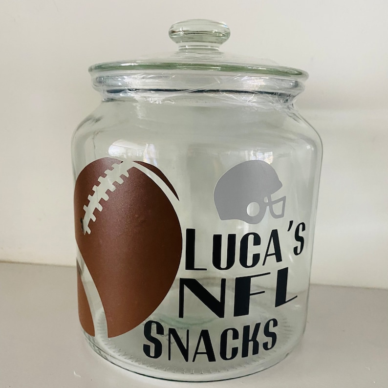 May include: A clear glass jar with a lid, decorated with a brown American football graphic and a grey helmet. The text "LUCA'S NFL SNACKS" is in black. The jar is designed for storing snacks.