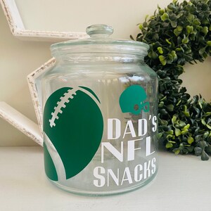 May include: A clear glass jar with a green football and helmet design. The jar has a white label that reads "Dad's NFL Snacks".