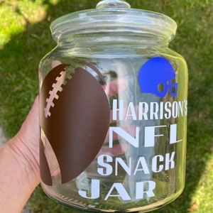 May include: A clear glass snack jar with a lid, featuring a brown American football graphic and a blue helmet. The jar has the text "Harrison's NFL Snack Jar" in white lettering.