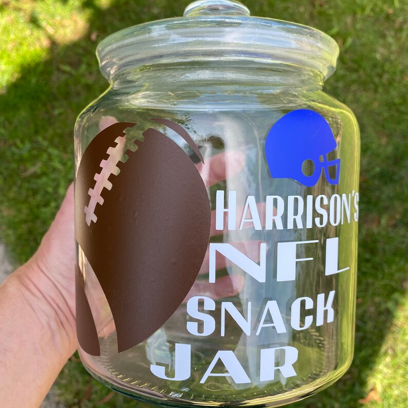 May include: A clear glass snack jar with a lid, featuring a brown American football graphic and a blue helmet. The jar has the text "Harrison's NFL Snack Jar" in white lettering.