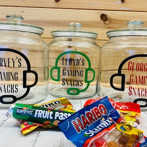 May include: Three clear glass jars with lids, each labeled "Gaming Snacks" with a name and a headset graphic in black, green, and red. Candy wrappers are scattered in front of the jars, including Haribo and Rowntrees.