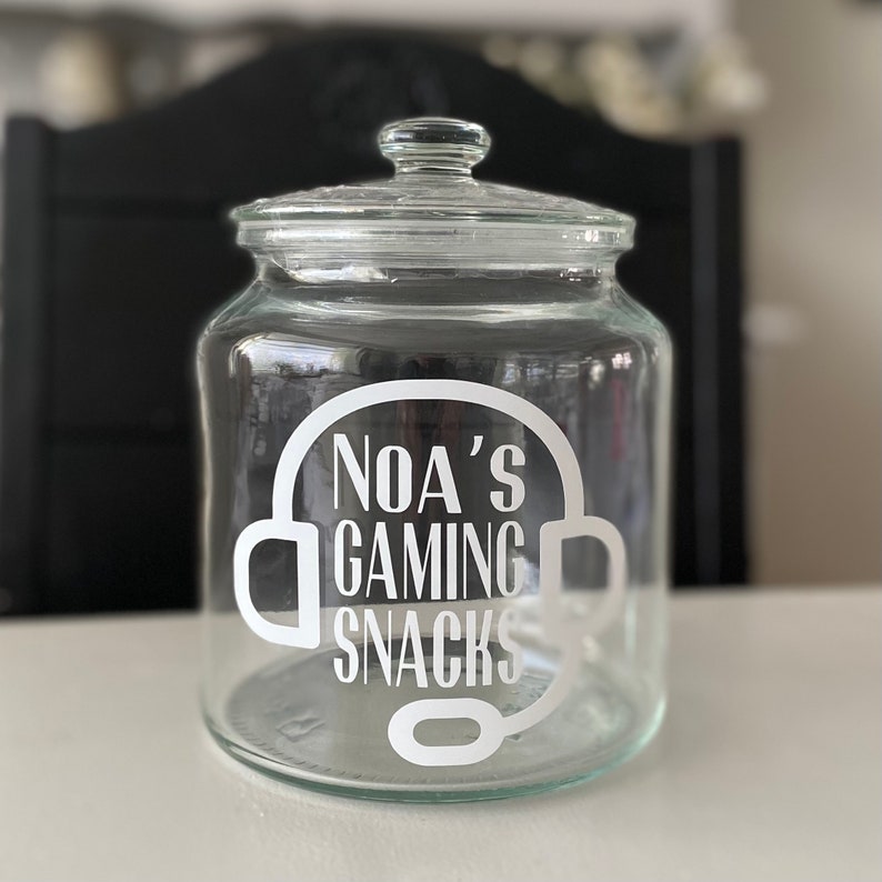May include: A clear glass jar with a lid, featuring the text "NOA'S GAMING SNACKS" in white, framed by a headset graphic. The jar is designed for storing snacks and is suitable for a gaming setup.