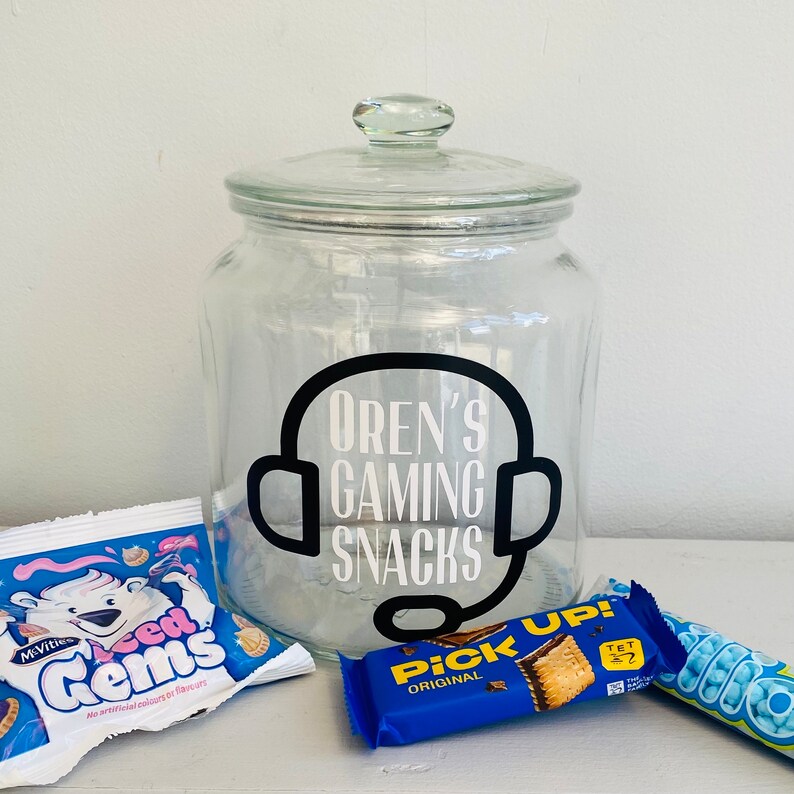 May include: A clear glass jar with a lid, labeled "Oren's Gaming Snacks" in white text with a black headset graphic. The jar is surrounded by snack packages, including a blue "Pick Up!" bar and a "McVities Iced Gems" bag.