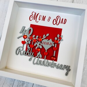 May include: A framed Ruby Anniversary gift. The white frame surrounds a red square with the words "Mum & Dad" and "Ruby Anniversary" in gray script. The number "40" and the word "Ruby" are in gray script. Red heart and gem accents are included.
