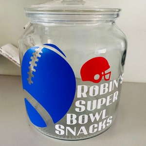 May include: A clear glass jar with a lid, decorated with a blue American football and a red helmet. The jar is labelled "Robin's Super Bowl Snacks" in white, ideal for storing snacks for a football game.