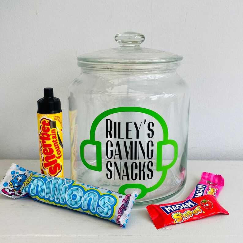 May include: A clear glass jar with a lid, labeled "Riley's Gaming Snacks" in green. The jar is surrounded by various candies, including a yellow Sherbet Fountain, blue Millions, and red and pink Maoam Stripes.