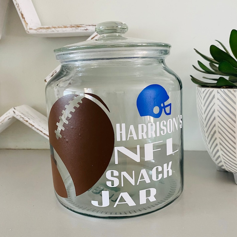May include: A clear glass snack jar with a lid, featuring a brown football graphic and a blue football helmet. The jar has the text "Harrison's NFL Snack Jar" in white. Ideal for storing snacks.