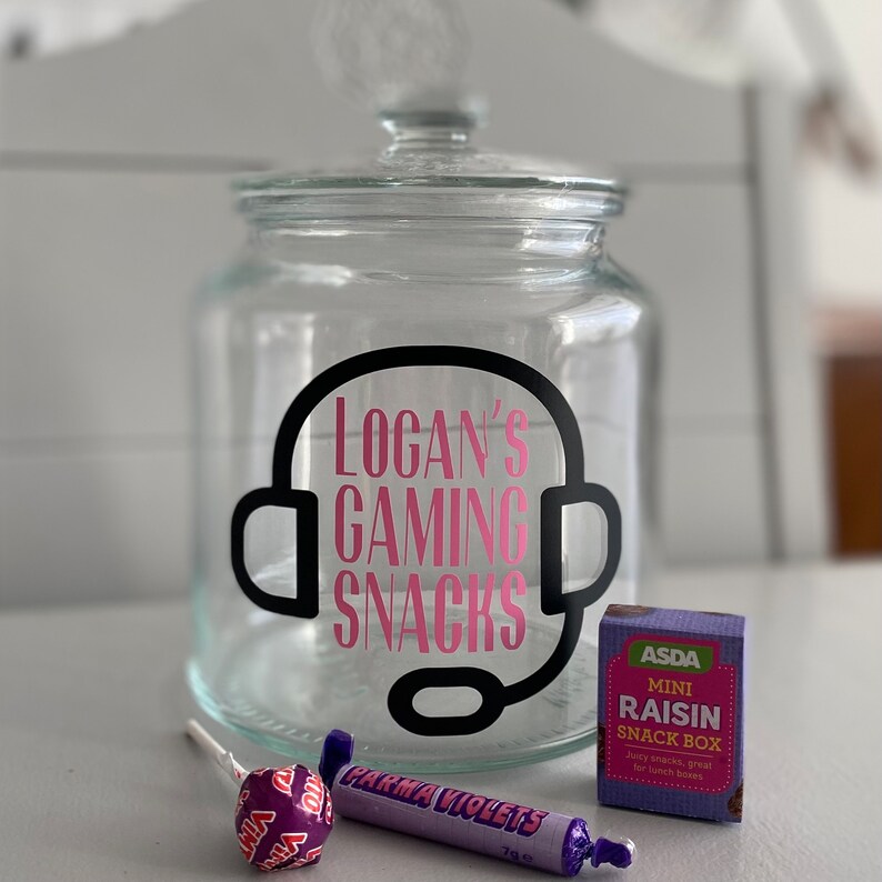 May include: A clear glass jar with a lid, featuring the text "Logan's Gaming Snacks" in pink, framed by a black headset graphic. Accompanied by a lollipop, a Parma Violets candy, and a mini raisin snack box.