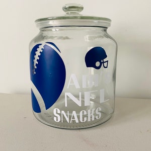 May include: A clear glass jar with a lid, decorated with a blue football and helmet graphic. White text reads "ADULT NFL SNACKS". The jar is designed for storing snacks.