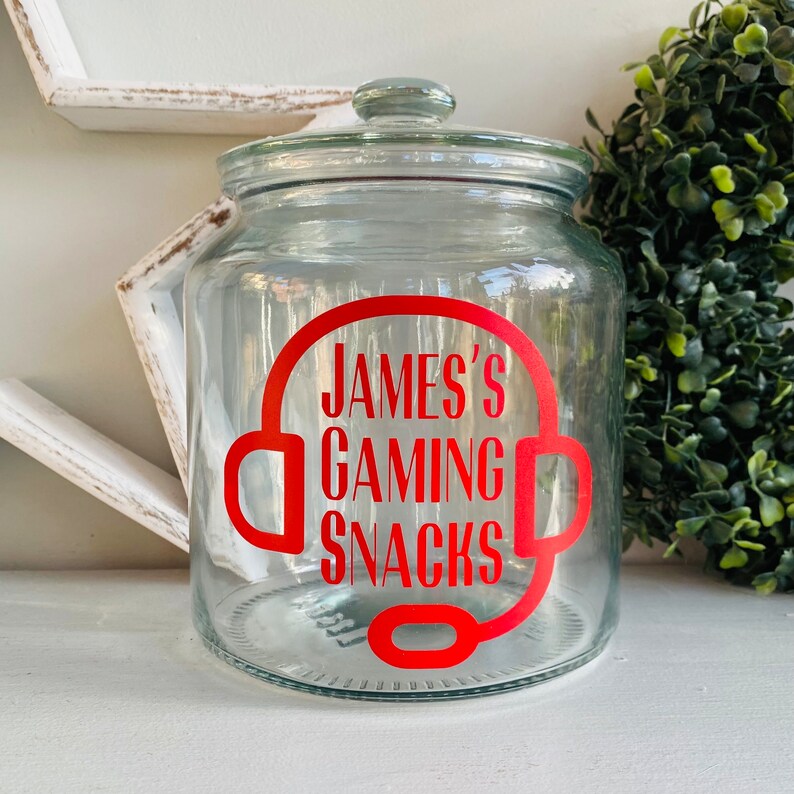 May include: A clear glass jar with a lid, featuring a red graphic of a headset and the text "JAMES'S GAMING SNACKS." The jar is designed for storing snacks and has a wide opening. The jar is sitting on a white surface.