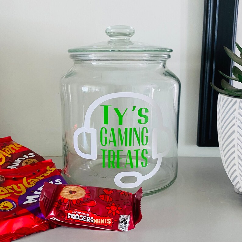 May include: A clear glass jar with a lid, labeled "Ty's Gaming Treats" in green and white. The jar is decorated with a headset graphic. Several packages of cookies are in front of the jar.