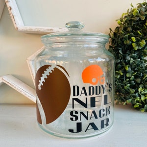 May include: A clear glass jar with a lid, decorated with a brown football, an orange football helmet, and the text "Daddy's NFL Snack Jar".