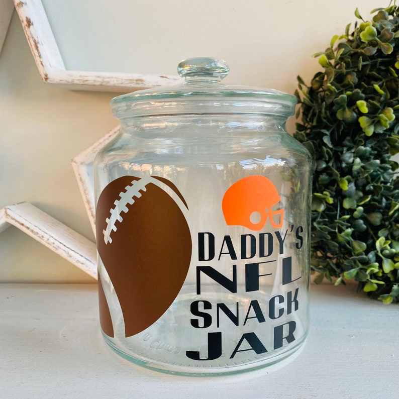 May include: A clear glass jar with a lid, decorated with a brown football, an orange football helmet, and the text "Daddy's NFL Snack Jar".