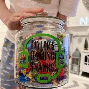 May include: A clear glass jar filled with colorful wrapped candies. The jar has a lid and a green decal that reads "Taylor's Gaming Snacks" with a headset design. The jar is held up by a person.
